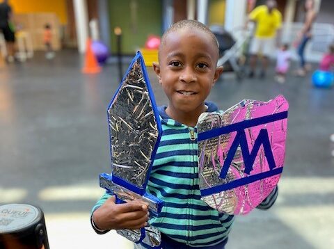 Child holding a homemade arts project, a cardboard sword and shield.